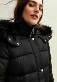 Czarna kurtka puchowa z kapturem wykończonym futrem, z zapięciem na zamek błyskawiczny i napy. Pikowana tekstura dodaje wymiaru. Srebrne akcenty metalowe.