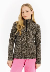 Blond pige iført en leopardmønstret langærmet zip-up trøje og lyserøde bukser, stående foran en hvid baggrund.