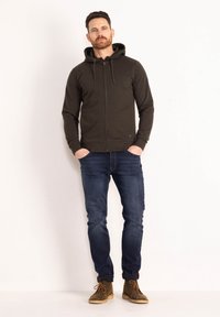 Felpa zip con cappuccio in verde scuro, abbinata a jeans blu scuro e stivaletti in suede marrone, in piedi su uno sfondo bianco.