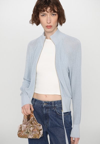Chaqueta de punto azul claro con cierre de cremallera, cuello acanalado y detalles plisados. Lleva una camiseta corta blanca y la combina con jeans oscuros, sosteniendo un pequeño bolso bordado.