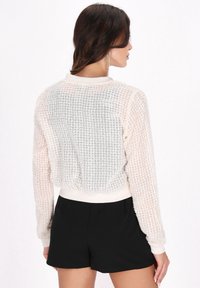 Leichter, semi-transparenter creamfarbener Cardigan mit einem strukturierten Karomuster; gerippte Bündchen und Saum; lässige Passform, kombiniert mit schwarzen Shorts.