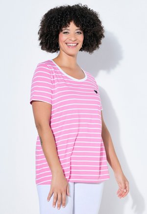 Femme souriante aux cheveux bouclés, portant une chemise rayée rose et blanche ainsi qu'un pantalon blanc, debout devant un fond blanc uni.