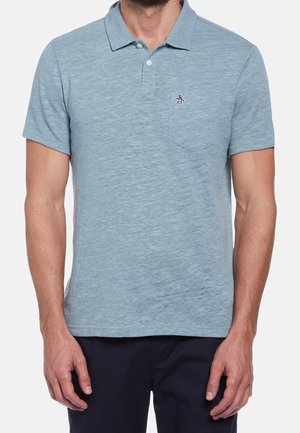 Mann trägt hellblaues kurzärmeliges Poloshirt mit kleinem Pinguin-Logo auf der Tasche und dunkle Hose, steht vor einfarbigem Hintergrund.