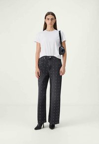 Mulher vestindo uma t-shirt branca, jeans pretos de cintura alta com bolinhas brancas, botas de salto pretas e uma pequena bolsa preta de ombro em fundo claro.