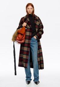 Mujer con cabello rojo que lleva un abrigo largo de cuadros rojos, verdes y negros, suéter negro, jeans azules, zapatos negros puntiagudos y lleva un bolso grande marrón.