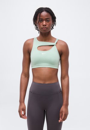 Atletisk kvinne med korte dreadlocks som har på seg en mintgrønn asymmetrisk sports-BH og høytlivs mørkegrå leggings, vendt fremover.