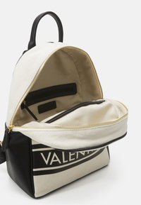 Mochila de lona con acentos de cuero negro, cremallera dorada y un bolsillo interior. Presenta un espacioso interior beige y un destacado estampado de logo.