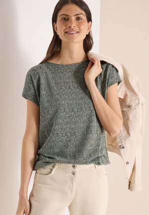 Femme souriante portant une chemise à manches courtes verte à motifs et un pantalon beige, tenant une veste beige sur son épaule.