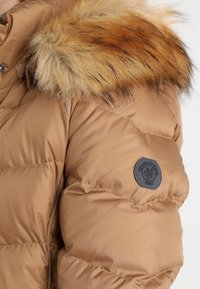 Chaqueta acolchada en nylon beige con textura de acolchado vertical, que incluye una capucha de piel sintética removible en tonos marrones y un parche con el logotipo en la manga.