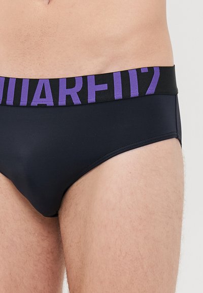 Nahaufnahme einer Person, die schwarze Unterwäsche mit einem breiten schwarzen Bund trägt, der große lila Buchstaben zeigt.