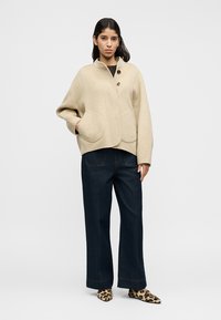 Μπεζ μάλλινο σακάκι με δομημένο σχεδιασμό, στρογγυλεμένο γιακά, δύο μεγάλα τσέπες και τρία κουμπιά. Συνδυάζεται με σκούρο τζιν παντελόνι φαρδιάς γραμμής.