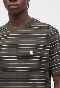 Άνδρας που φορά κοντομάνικο T-shirt με ρίγες σε χακί, λευκό, μαύρο και τιρκουάζ χρώμα με λευκό κεντημένο λογότυπο στο στήθος πάνω σε λευκό φόντο.