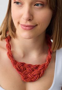 Vrouw die een ingewikkeld gevlochten koraal en rode kralenketting draagt met een witte top tegen een neutrale achtergrond.
