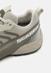 Lichtgrijs sportshoe met bovenkant van gaas, donkergroene accenten, witte "SCARPA" logo, gestructureerde zool en versterkte hieltab.