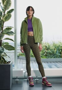 Grøn fleecejakke over en lilla cropped top, parret med oliven leggings med logo-print og pink- og grønne sneakers. Indendørs miljø.