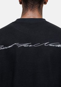 Schwarzes, texturiertes T-Shirt mit Rundhalsausschnitt und einem weißen, gestickten Spruch "Good For Nothing" auf der Rückseite.