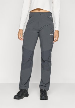 Inimene, kes kannab tumedat halli matkaretuusid tugevdatud põlveplokkidega ja musti matkajalatsid, valge ühevärvilise tausta ees.