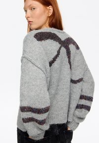 Młoda kobieta z rudymi włosami, ubrana w szary, dziergany sweter z czarnymi, błyszczącymi paskami na rękawach i plecach, patrząca przez ramię.