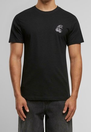 Mann trägt ein schwarzes T-Shirt mit kurzen Ärmeln und einem kleinen weißen Kirsch-Design auf der linken Brust, kombiniert mit dunkelgrauer Hose.
