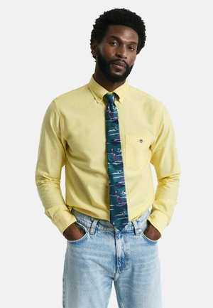 Chemise jaune à manches longues avec col à boutons, associée à une cravate à motifs présentant des vagues bleues, roses et grises. Portée avec un jean bleu clair.