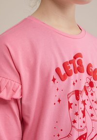 Rožinė medvilninė marškinė su trumpomis, pūstomis rankovėmis. Kitoje pusėje yra didelis raudonas tekstas "EIKIME" ir grafika su batais, žvaigždėmis bei gėlių raštu.