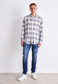 Camisa de manga larga con botones en un diseño a cuadros azul y crema, combinada con jeans azules y zapatillas blancas. Textura suave y un corte relajado.