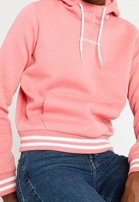 Sudadera rosa claro con un bolsillo frontal, cordones blancos y puños acanalados con rayas blancas en el dobladillo. Textura de tela suave.