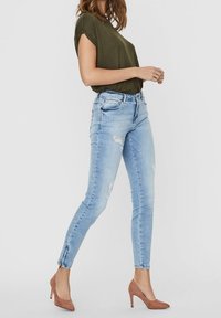 Světlé modré skinny džíny se zeslabenými detaily a rozparky u kotníků, v kombinaci s tmavě zeleným tričkem a béžovými vysokými podpatky.