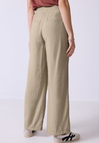 Beige wijde broek gemaakt van een textuurstof met een elastische tailleband en twee achterzakken, gecombineerd met witte schoenen met zwarte accenten.