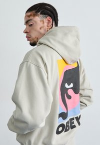 Felpa con cappuccio beige caratterizzata da una grande stampa grafica con un design nero e vivaci colori di sfondo, che mostra la parola "OBEY."