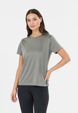 Kvinde står med hånden på hoften iført en ensfarvet grå T-shirt med korte ærmer og sorte leggings mod en hvid baggrund.