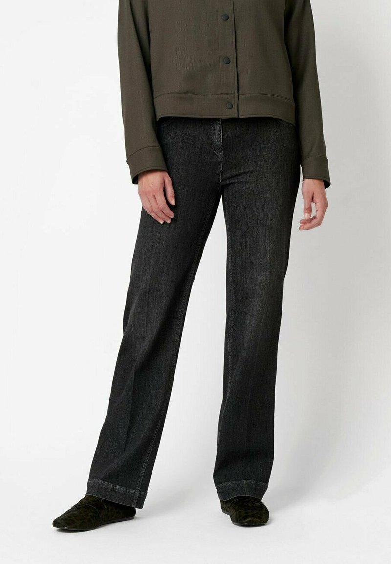 Schwarze Jeans mit geradem Schnitt, kombiniert mit einer dunkelgrünen, knöpfbaren Jacke, die ein strukturiertes Design und einen runden Kragen aufweist.