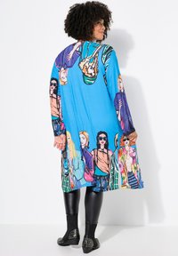 Long, blue dress featuring colorful, illustrated women in various outfits. Made of smooth fabric with a relaxed fit and long sleeves, paired with black leggings and studded ankle boots.
