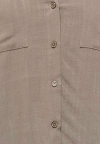 Chemise en tissu taupe avec quatre boutons beiges alignés au centre et poches à la poitrine cousues horizontalement de chaque côté.