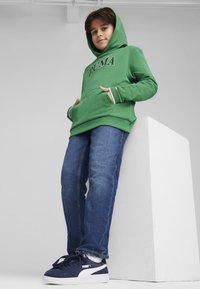 Felpa verde con cappuccio con logo "PUMA", abbinata a jeans di denim blu e sneakers blu scuro con dettagli bianchi.