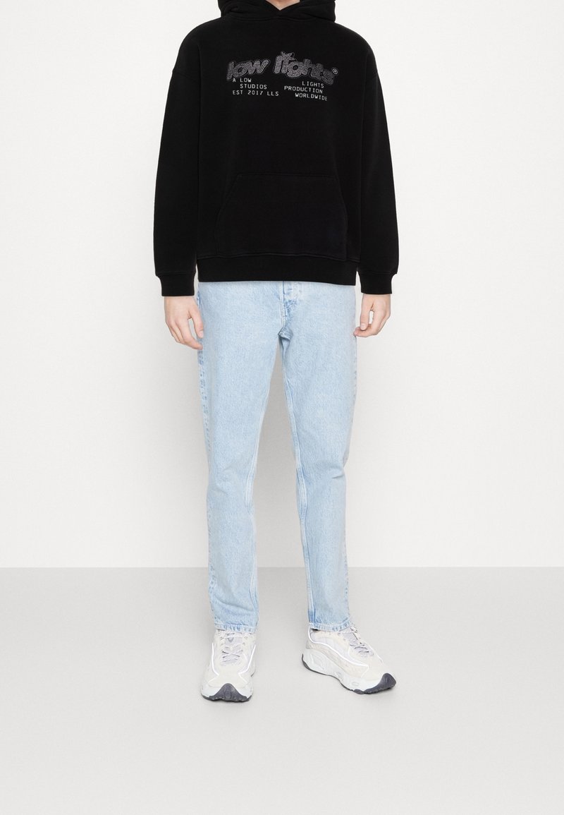 Sweat à capuche noir avec poche avant et logo, assorti à un jean slim bleu clair et des baskets blanches avec des accents gris.