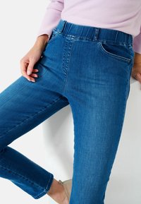 Jean en denim bleu avec une texture lisse, taille mi-haute à bande élastique, et deux poches avant. Design ajusté se rétrécissant au niveau de la cheville.