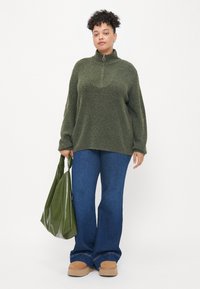 Púrpura de punto verde con cuello con cremallera, jeans de mezclilla azules de corte acampanado y zapatos de lona beige. Sosteniendo una bolsa de mano verde brillante.