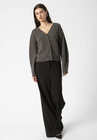 Un model merge cu încredere într-un cardigan gri și pantaloni larghi negri, pe un fundal alb simplu.
