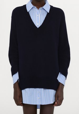 Personne portant une robe-chemise boutonnée à rayures bleues et blanches sous un pull ample noir à encolure en V.