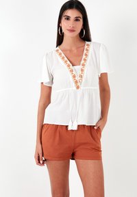 Witte blouse met oranje bloemenborduursel langs de V-hals; korte mouwen. Gecombineerd met roestkleurige shorts. Gelaagd stof.