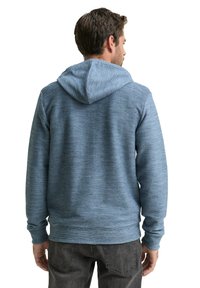 Mężczyzna zwrócony plecami, ubrany w niebieską, fakturowaną bluzę z kapturem i ciemnoszare dżinsy na białym tle.