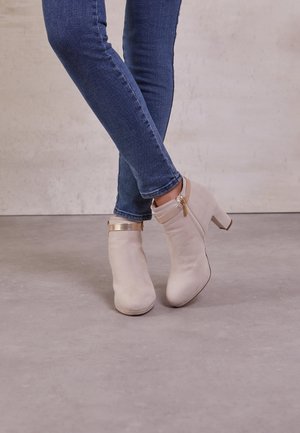 Ben krydsede, iført tætsiddende blå jeans og beige ankelstøvler med blokhæle og gyldne lynlåse på en glat grå gulv.