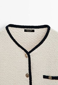 Cardigan texturé crème avec bordure noire, boutons décoratifs dorés et petit détail de poche, étiqueté Made in Turkey.