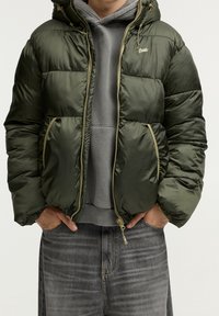 Chaqueta acolchada en verde oscuro, con capucha, cierre de cremallera en la parte delantera y puños elásticos. Dos bolsillos laterales con detalles en amarillo y diseño acolchado.