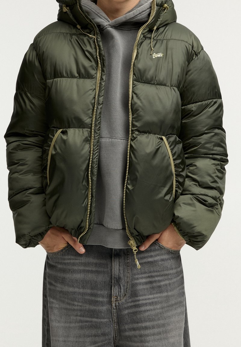 Chaqueta acolchada en verde oscuro, con capucha, cierre de cremallera en la parte delantera y puños elásticos. Dos bolsillos laterales con detalles en amarillo y diseño acolchado.