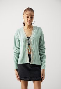 Mintgrønn langermet cardigan med knyttefront, blomsterbroderi langs åpningen og en cropped design, kombineres med et svart denimskjørt.