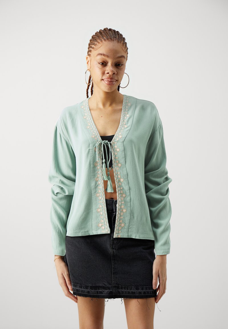 Mintgrønn langermet cardigan med knyttefront, blomsterbroderi langs åpningen og en cropped design, kombineres med et svart denimskjørt.