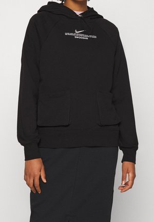 Personne portant un sweat à capuche noir avec des poches avant et un texte blanc imprimé indiquant "SWOOSH" sur la poitrine.