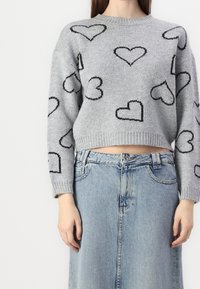 Šedý cropped svetr s černě obrysy srdcí, ve spojení s džínovou sukní s vysokým pasem v světle modré barvě, nošený stojící osobou.